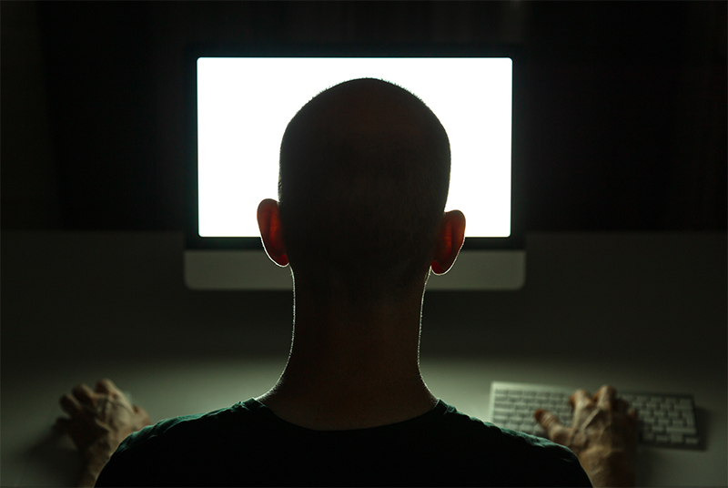 A silhouetted hacker sits in front of a computer screen in a dark room.
