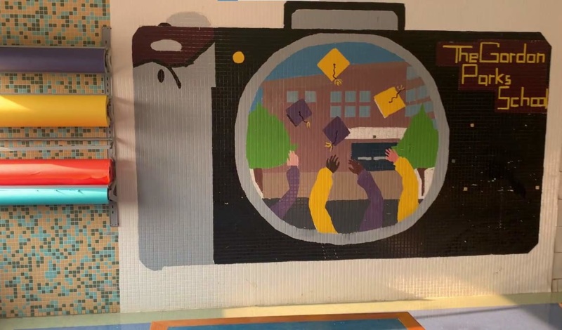 A photo of a painted mural on the wall at The Gordon Parks School for Inquisitive Minds. The mural depicts a black camera, and in the camera lens is an image of students throwing their graduation caps in the air in front of the school. The words painted in the top right side of the camera reads 
