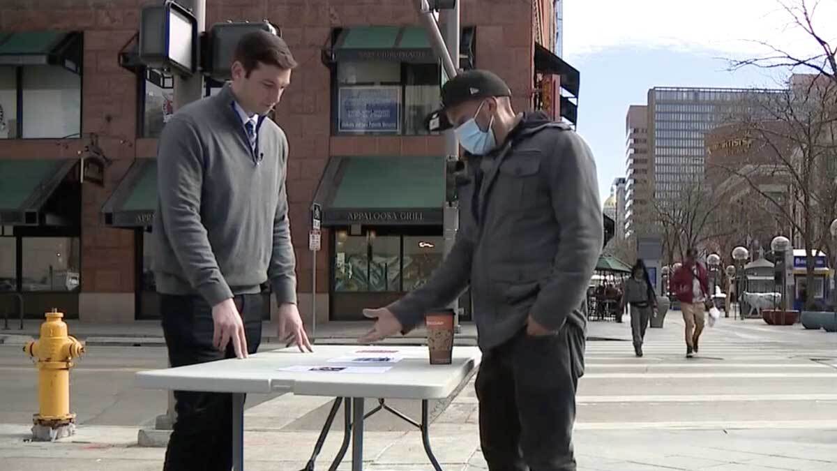 Two men stand out on a street corner, on either side of a folding table, examining and gesturing toward images taped to the table.