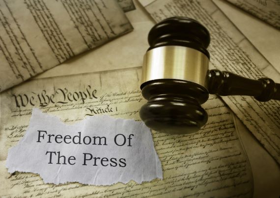 An illustration of the U.S. Constitution with a gavel on top along with a torn white piece of paper with “Freedom Of The Press” typed on it.