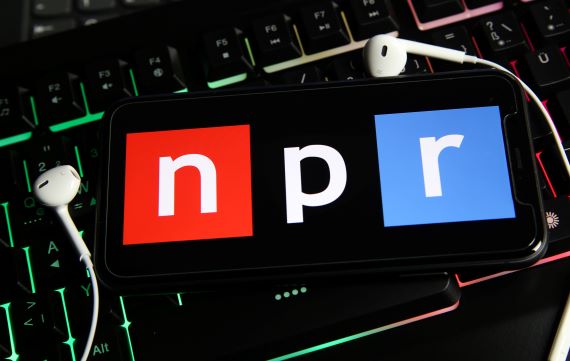 The NPR logo on a smartphone screen rests above a black keyboard with backlit keys and white earbuds.