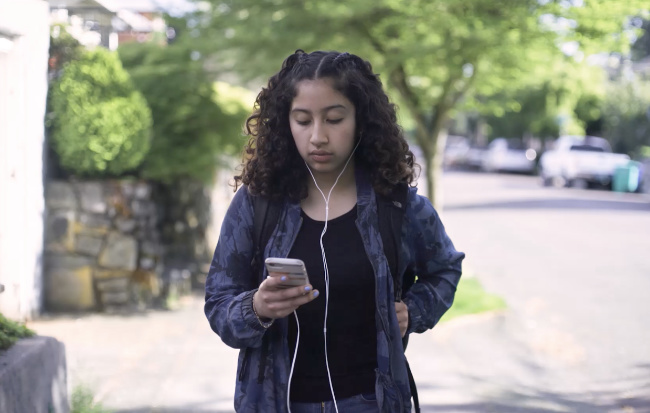 A girl wearing headphones and a backpack looks down at a smartphone while standing outside.