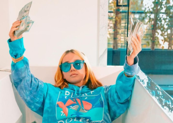Teenaged rapper Lil Tay sits in a dry bathtub wearing a blue sweatshirt and blue sunglasses while holding bundles of cash in her hands.