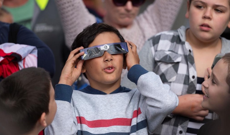A young boy looks upwards while holding solar eclipse glasses to his eyes. Other adults and children are gathered around him.