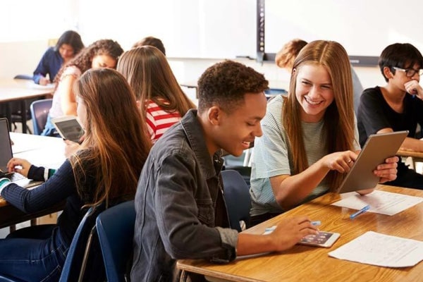 An image of several teenaged students sitting at their desks in a classroom with laptops, paper and a device out.