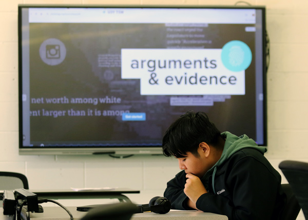 A student looks down at a worksheet during class. Behind him is the classroom video monitor displaying Checkology’s “Arguments and Evidence” lesson on screen.