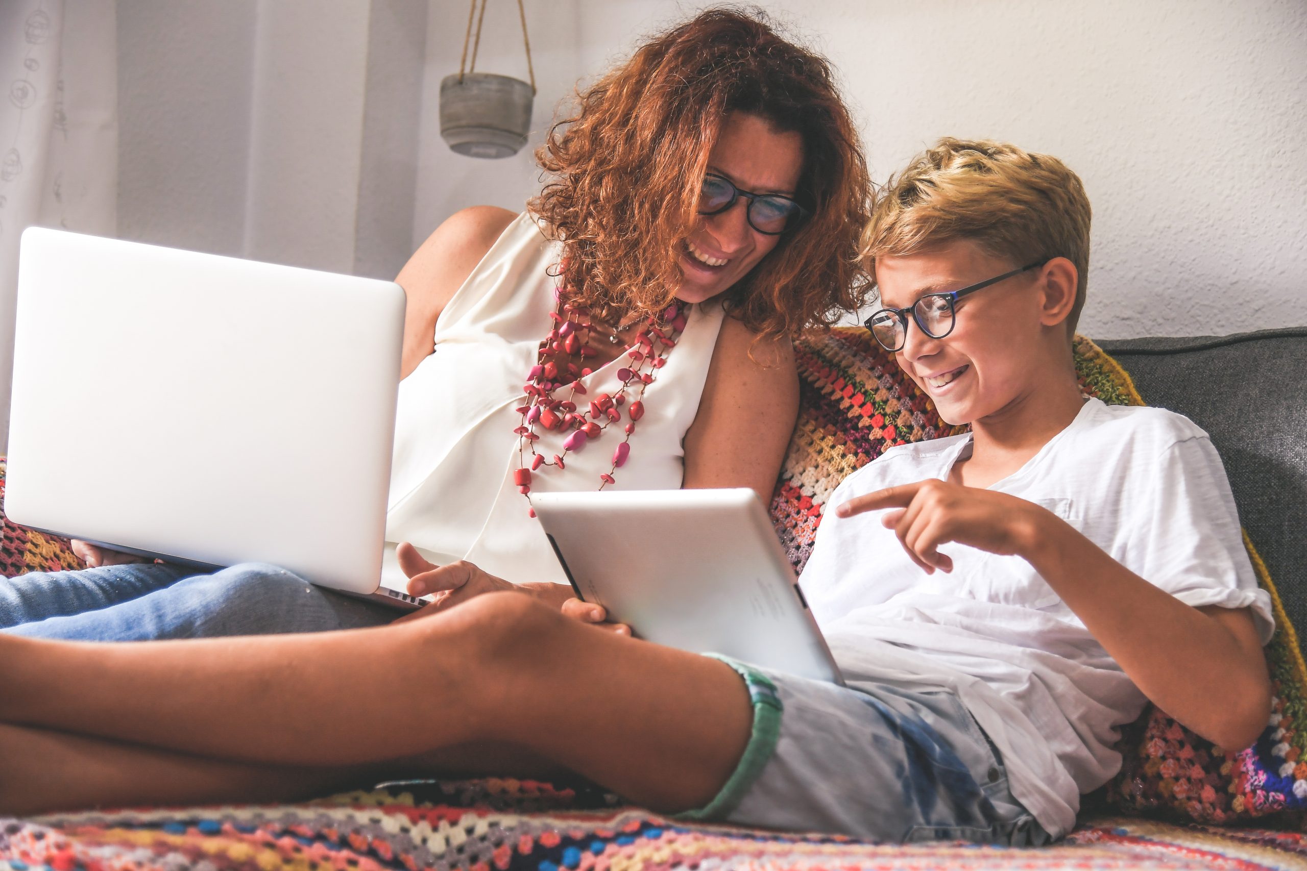 Red haired mom sitting with blond haired son, looking over his should at his tablet.