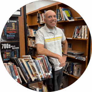 Photo of Philip Lensi smiling at the camera in a library leaning against a stack of books.