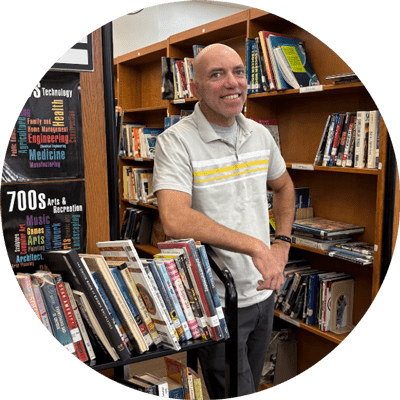 Photo of Philip Lensi smiling at the camera in a library leaning against a stack of books.