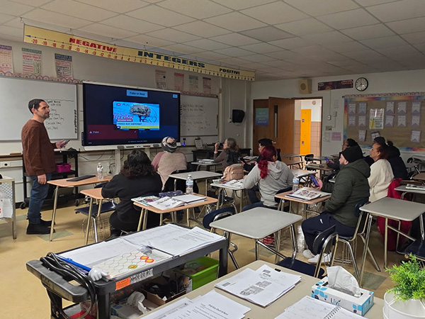 Alex Luciano, who teaches English as a second language to adults in Central Islip, N.Y., stands at the front of his classroom while students look at an image on a large screen and then describe what they see.