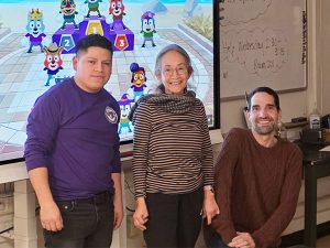 Alex Luciano, who teaches English as a second language to adults in Central Islip is show to the right of two of his students in his classroom. Behind them a cartoon image, which he uses as a teaching tool, is displayed on a large screen.