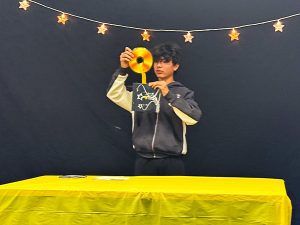 Student holds up a gold CD from a black case decorated with stars during his class magic show. String lights with stars hang in the background.