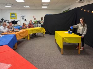 Student stacks a set of blue cups on a yellow table during her class magic show. A panel of school administrators and educators watch from the audience.