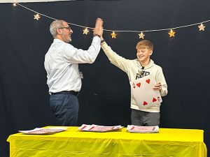 A school administrator and a student share a high-five during his class magic show. The student holds a large playing card, with a stack of cards on the yellow table in front of them. String lights with stars hang in the background.
