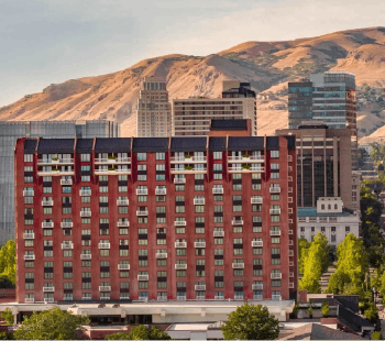 Red brick mid-rise apartment building in a city, with hills and modern glass buildings in the background behind it.
