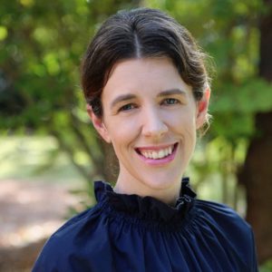 Smiling woman with dark hair in a navy blouse outdoors in a green park setting.
