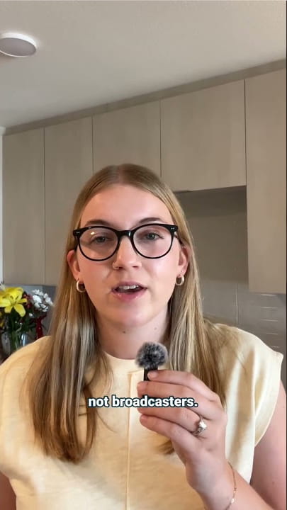 Woman wearing glasses speaks to the camera in a kitchen, holding a small fluffy microphone; on-screen caption reads 'not broadcasters'.