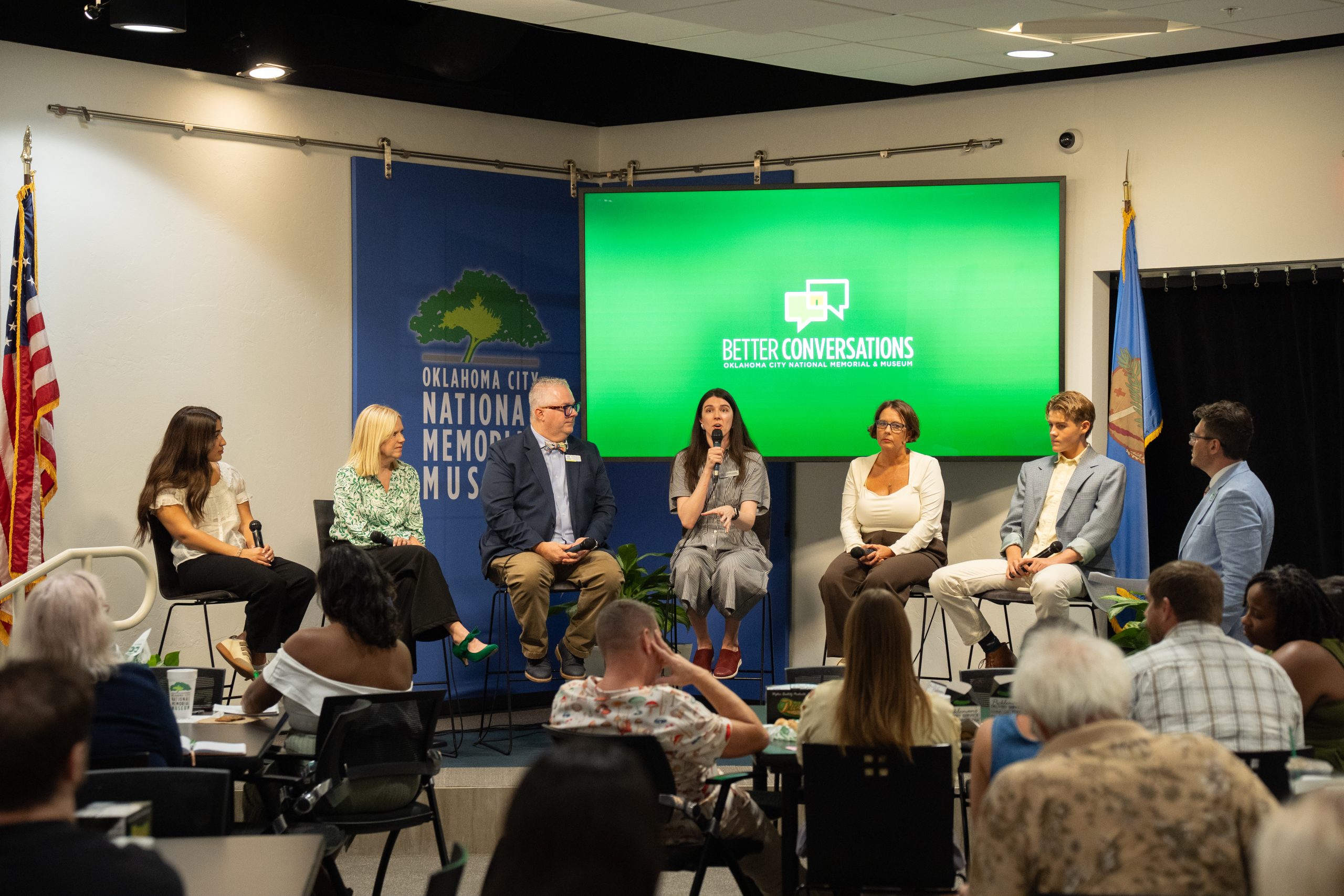 Six panel members sit in a semicircle on a stage in front of a large screen that says, 